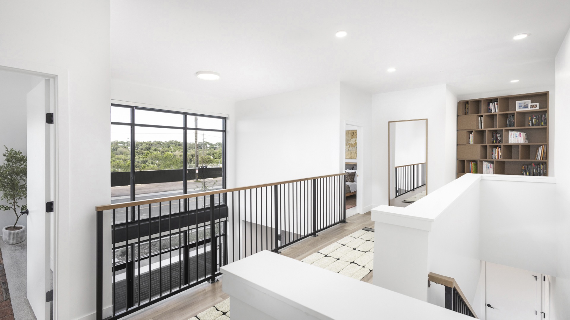 Bright and modern upper-level hallway with expansive windows, showcasing lush greenery and featuring a sleek railing and built-in bookshelf.	